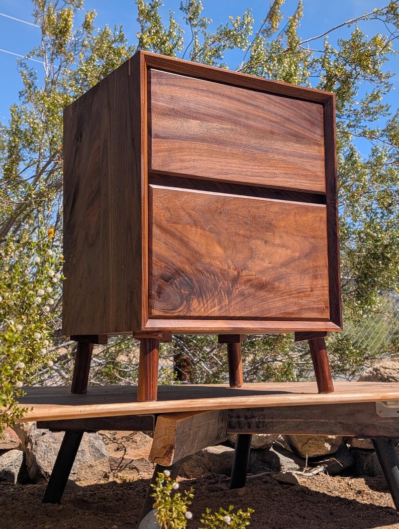 Pu&ograve; includere: Un comodino in legno marrone scuro con due cassetti sovrapposti caratterizzati da una venatura del legno naturale visibile. Il mobile &egrave; sostenuto da quattro gambe cilindriche corte e presenta una struttura rettangolare con bordi dritti.