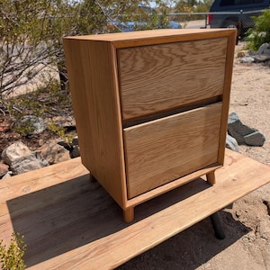 Pu&ograve; includere: Un comodino in legno marrone chiaro dotato di due cassetti sovrapposti con una venatura del legno naturale visibile. Il mobile &egrave; costruito con una struttura rettangolare ed &egrave; sostenuto da quattro gambe cilindriche corte in legno alla base.