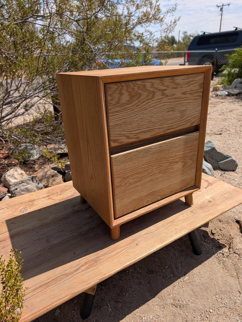 Pu&ograve; includere: Un comodino in legno marrone chiaro dotato di due cassetti sovrapposti con una venatura del legno naturale visibile. Il mobile &egrave; costruito con una struttura rettangolare ed &egrave; sostenuto da quattro gambe cilindriche corte in legno alla base.