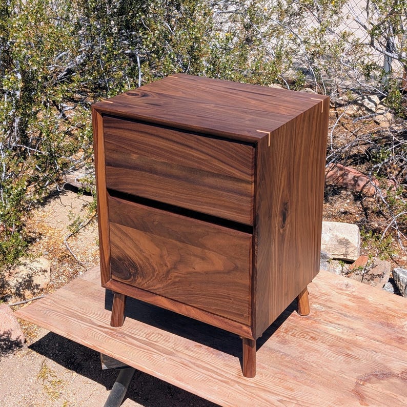 Pu&ograve; includere: Un comodino in legno marrone scuro con due cassetti sovrapposti e una venatura del legno visibile. Il mobile &egrave; costruito con linee dritte e pulite ed &egrave; sostenuto da quattro gambe corte e affusolate. Sono visibili piccoli accenti in legno chiaro.