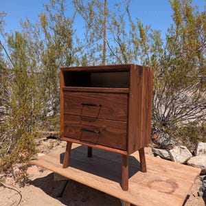 Può includere: Un comodino in legno marrone scuro dotato di un ripiano superiore aperto e due cassetti con sottili maniglie in metallo nero. Il mobile presenta venature del legno visibili ed è sostenuto da quattro gambe in legno affusolate.