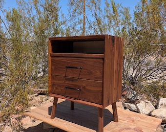 May include: A dark brown wooden bedside cabinet featuring an open top shelf and two drawers with thin black metal handles. The piece is constructed with visible wood grain and is supported by four tapered wooden legs extending from the base.