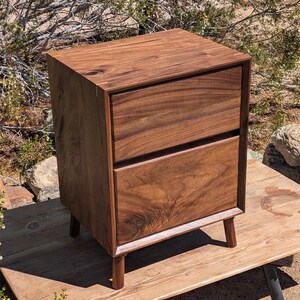 Pu&ograve; includere: Un comodino rettangolare realizzato in legno di noce marrone scuro con una venatura naturale visibile. Il mobile presenta due cassetti sovrapposti con frontali piatti ed &egrave; sostenuto da quattro gambe cilindriche corte in legno.