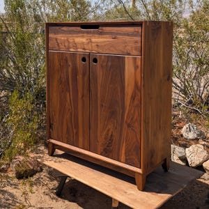Può includere: Un mobile alto in legno marrone scuro con due ante e un cassetto. Il mobile ha un design semplice con fori circolari per le maniglie. È posto su una piattaforma di legno e ha quattro gambe corte. Misura circa 100 cm di altezza.
