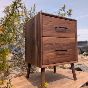 May include: A two-drawer wooden nightstand with a warm brown finish. The nightstand has black handles and angled legs. The nightstand is approximately 24 inches tall, 18 inches wide, and 16 inches deep.