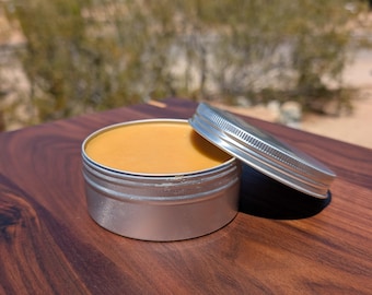 May include: Open silver tin containing a light brown, creamy substance. The lid is ajar, showing the product. The tin rests on a dark brown wooden surface, with a blurred natural background.