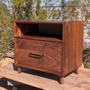 May include: A walnut nightstand with a drawer and open shelf. The nightstand has a dark finish, black drawer pull, and four short legs. The nightstand is approximately 24 inches wide, 18 inches deep, and 24 inches tall.