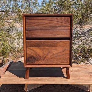 May include: A rectangular wooden nightstand featuring two stacked drawers with a visible dark brown wood grain pattern. The piece is constructed with a solid frame and is supported by four short, tapered legs extending from the bottom corners.