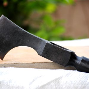 Könnte beinhalten: Ein schwarzer, handgeschmiedeter Tomahawk mit gebogener Klinge und Hammerkopf am gegenüberliegenden Ende. Der Axtkopf hat ein Loch in der Nähe des Hammers. Der Tomahawk liegt auf einer hellen Oberfläche.