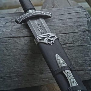 Peut inclure: Épée viking avec un manche en bois foncé et une lame argentée. La lame porte des runes gravées. L'épée est dans un fourreau noir avec des détails argentés et une sangle décorative. Image détaillée.