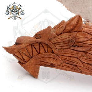 May include: A wooden carving of a dragon's head with detailed scales and an open mouth, showing sharp teeth. The carving is made of a light brown wood and has a natural finish.