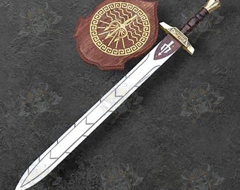May include: A decorative sword with a silver blade and a brown and gold handle, resting beside a wooden shield with a gold emblem. The sword has a detailed design on the blade and handle. The shield is a dark brown color.