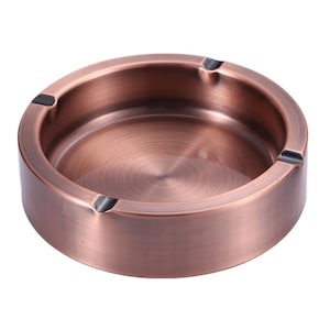 May include: A round, copper-colored ashtray with a brushed metal finish. The ashtray has a low profile with a circular design and three small notches for resting cigarettes. The interior has a concentric circle pattern.
