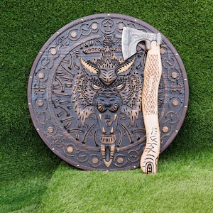 Op de afbeelding: Een houten schild met een gesneden wolfskopontwerp en een decoratieve bijl met een houten handvat. Het schild is bruin en heeft een leren riem. De bijl heeft een zilveren blad en een houten handvat met ingewikkelde houtsnijwerk.