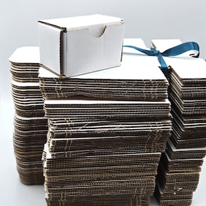 May include: Stacks of small, white cardboard boxes with a natural brown corrugated interior. One box sits atop a stack, with a small cutout on the side. A blue ribbon is tied around one stack. Ideal for packaging small items.