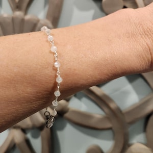 Könnte beinhalten: Ein weißes Perlenarmband mit einem silbernen Verschluss. Das Armband befindet sich an einem Handgelenk.