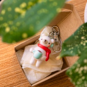 Peut inclure: Un porte-cl&eacute;s en c&eacute;ramique blanche en forme d'ours tenant une tasse bleue, portant une &eacute;charpe rouge et un bandeau blanc. Le porte-cl&eacute;s est dans une bo&icirc;te brune avec du papier de soie blanc.