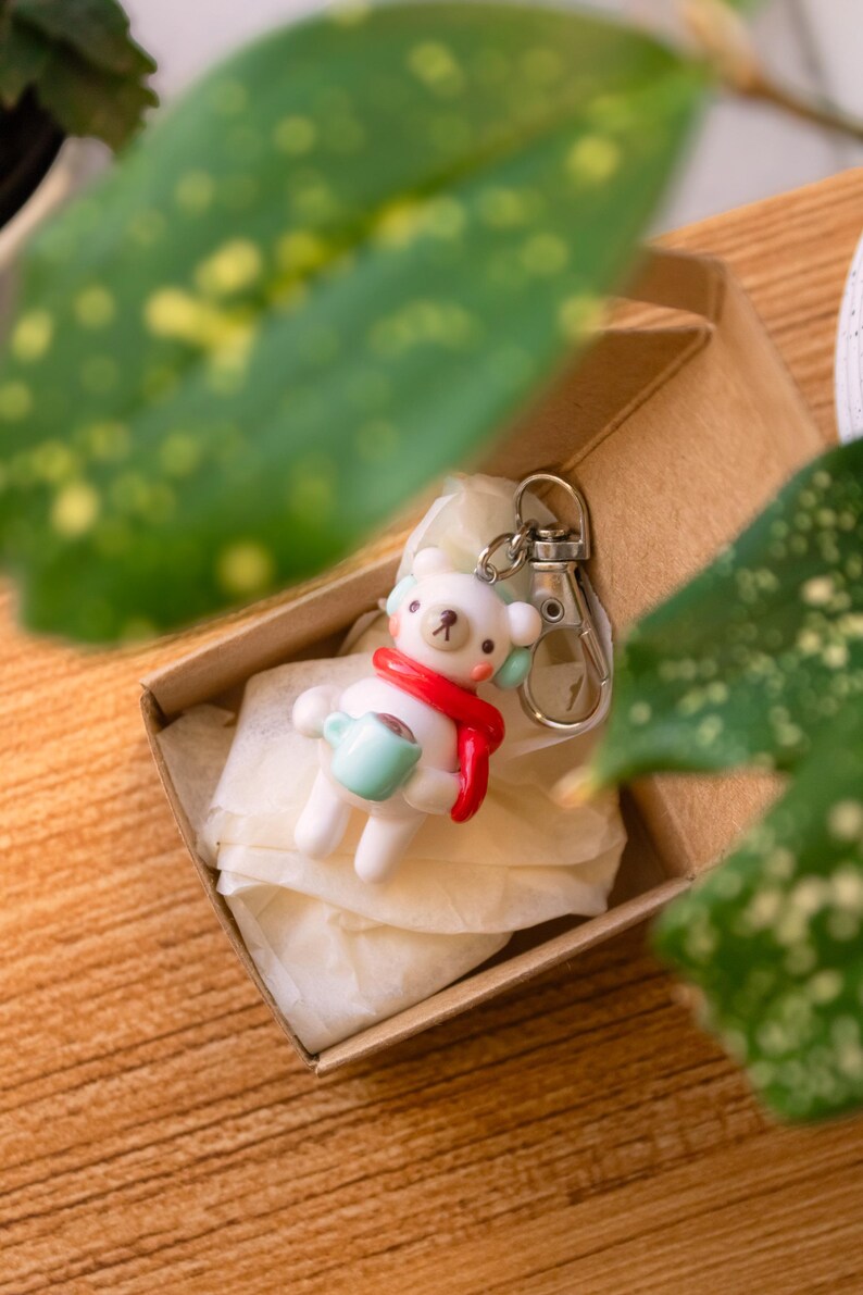 Peut inclure: Un porte-cl&eacute;s en c&eacute;ramique blanche en forme d'ours tenant une tasse bleue, portant une &eacute;charpe rouge et un bandeau blanc. Le porte-cl&eacute;s est dans une bo&icirc;te brune avec du papier de soie blanc.