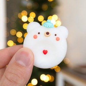 May include: A white, bear-shaped decorative item with a light blue hat, small brown nose, and red heart. The bear has pink cheeks and small black eyes. The item is held in a person's hand against a blurred background of golden lights.