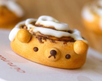 May include: A cinnamon roll shaped like a bear with white frosting and chocolate eyes. The bear is sitting on a white surface.