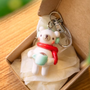 Peut inclure: Un porte-cl&eacute;s en c&eacute;ramique blanche en forme d'ours portant une &eacute;charpe rouge et un casque vert, tenant une tasse bleu clair avec une boisson brune &agrave; l'int&eacute;rieur. Le porte-cl&eacute;s est dans une bo&icirc;te brune avec du papier de soie blanc.