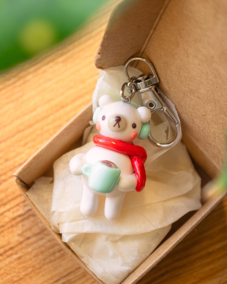 Peut inclure: Un porte-cl&eacute;s en c&eacute;ramique blanche en forme d'ours portant une &eacute;charpe rouge et un casque vert, tenant une tasse bleu clair avec une boisson brune &agrave; l'int&eacute;rieur. Le porte-cl&eacute;s est dans une bo&icirc;te brune avec du papier de soie blanc.