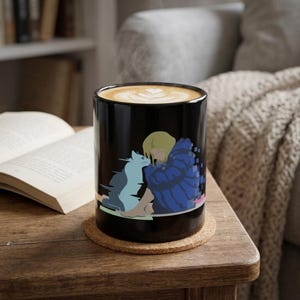 May include: A black ceramic mug filled with coffee featuring a latte art heart design. The exterior of the mug displays a graphic illustration of a blonde figure in a blue jacket sitting next to a blue cat, with pixelated edges on the jacket.