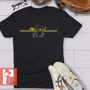 Op de afbeelding: Donkergrijs t-shirt met een geel en grijs grafisch ontwerp. Het ontwerp bevat een gestileerde "A" en geometrische vormen. Het shirt wordt getoond met witte sneakers, een bril en een bruine leren tas.