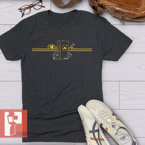 Op de afbeelding: Donkergrijs t-shirt met een geel grafisch ontwerp. Het ontwerp toont een gestileerde afbeelding met geometrische vormen en de letter "A". Het shirt heeft korte mouwen en is gemaakt van een zacht materiaal. Witte sneakers met oranje en marine accenten staan op de voorgrond.