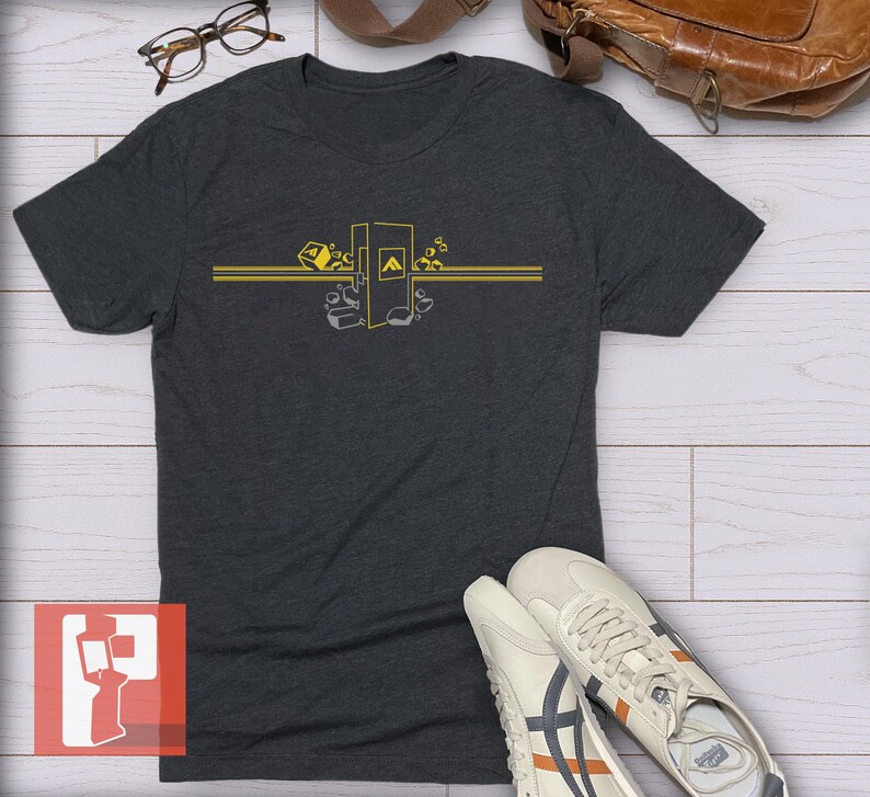 Op de afbeelding: Donkergrijs t-shirt met een geel grafisch ontwerp. Het ontwerp toont een gestileerde afbeelding met geometrische vormen en de letter "A". Het shirt heeft korte mouwen en is gemaakt van een zacht materiaal. Witte sneakers met oranje en marine accenten staan op de voorgrond.