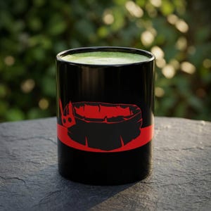 May include: A black ceramic mug filled with a vibrant green liquid. The exterior features a horizontal red band encircling the middle, with a stylised red graphic of a small, horned creature head positioned on the left side of the mug's surface.