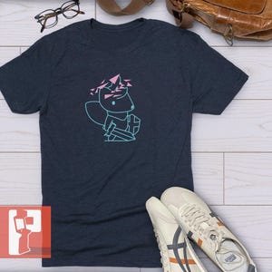 May include: A dark navy blue short-sleeve t-shirt featuring a light blue line art graphic of a small creature holding a sword and shield, with pink geometric shapes floating above its head. The design is centred on the chest of the garment.