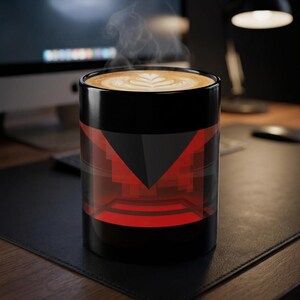 May include: A black ceramic mug filled with a hot latte featuring a white leaf-shaped foam design. The exterior of the mug displays a geometric pattern with a large dark triangle pointing downward over a vibrant red and black pixelated background.