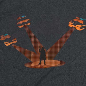 May include: Dark gray t-shirt featuring an orange graphic of a person in silhouette standing in a spotlight, with three orange vehicles above. The vehicles have blue accents. The spotlight beams have a dotted pattern.