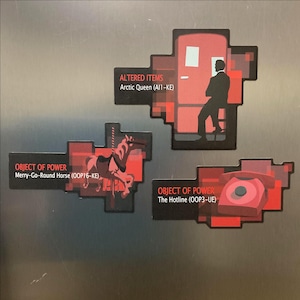May include: Three rectangular magnets with red and black pixelated backgrounds. One shows a silhouette of a person by a door labeled Altered Items Arctic Queen (AI1-KE). Another shows a carousel horse labeled Object of Power Merry-Go-Round Horse (OOP16-KE). The third shows a red telephone labeled Object of Power The Hotline (OOP3-UE).