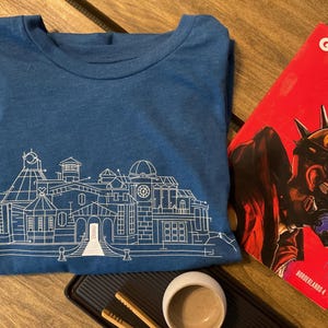 May include: A blue t-shirt with a white architectural line drawing. A red magazine with a character illustration and the word "GAME" is next to the shirt. A small white cup and a black tray are also in the image.