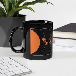 May include: Black ceramic mug with a white and orange design of a solar system. The mug has a large orange sun and smaller white planets and rings.