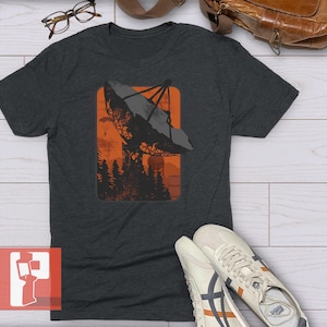 May include: Dark gray t-shirt featuring a graphic of a radio telescope in an orange rectangle, set against a forest silhouette. The tee is a crew neck and is paired with white sneakers with orange and navy accents.