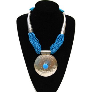 Puede incluir: Un collar de plata con un gran colgante redondo que presenta una piedra turquesa. El colgante está rodeado por una franja de cuentas de color azul turquesa.