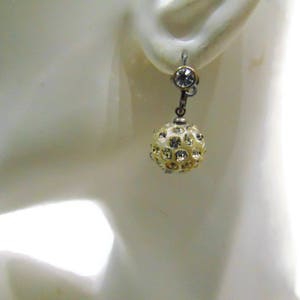 Könnte beinhalten: Ein einzelner Ohrring mit einer cremefarbenen, mit Strasssteinen besetzten Kugel. Der Ohrring ist oben mit einem kleinen, klaren Strassstein und einem silberfarbenen Metallverschluss befestigt. Schmuck.
