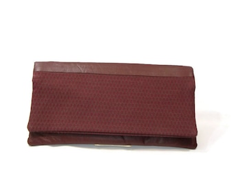 May include: A burgundy leather clutch with a textured fabric flap. The clutch has a silver clasp.