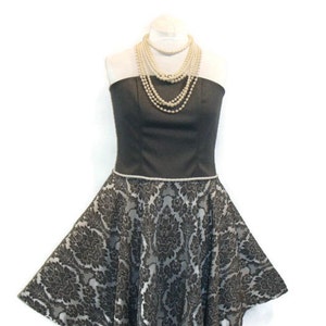 Könnte beinhalten: Ein trägerloses graues und weißes Kleid mit Damastmuster. Das Kleid hat ein tailliertes Oberteil und einen vollen Rock. Das Kleid ist mit einer Perlenkette verziert.