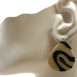 Könnte beinhalten: Ein Paar Ohrringe mit Tropfenform. Die Ohrringe sind aus beigefarbenem Material mit schwarzen und goldenen Spiralen.