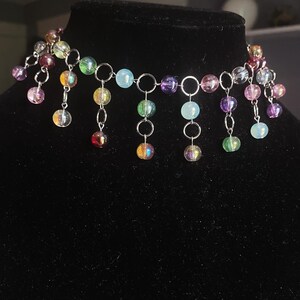 May include: A colorful beaded choker necklace. The necklace features a series of iridescent, round beads in various colors, including purple, green, yellow, and blue. The beads are connected by silver-toned metal rings and chains, creating a cascading effect.