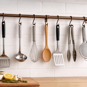 May include: A bronze-coloured metal kitchen rail mounted horizontally, featuring seven S-shaped hooks. Hanging from these hooks are a slotted turner, a ladle, a wire whisk, a wooden spoon, a second slotted turner, metal tongs, and a fine mesh strainer.
