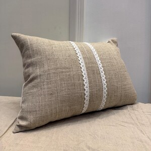 Puede incluir: Una almohada rectangular beige con dos tiras verticales de encaje blanco. La almohada tiene un aspecto de tela texturizada, y el encaje añade un elemento decorativo. La almohada está sobre una superficie beige.