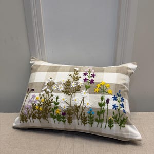 Puede incluir: Un cojín rectangular con un patrón a cuadros beige y blanco. Está bordado con un diseño floral colorido con margaritas blancas, flores moradas, amarillas y azules, y tallos verdes.