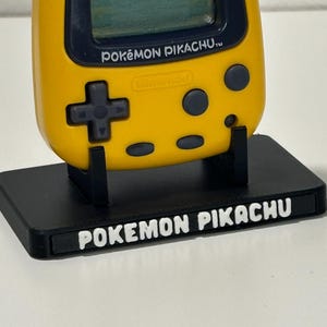 Peut inclure: Un appareil de jeu électronique portable jaune étiqueté POKEMON PIKACHU avec une croix directionnelle et des boutons gris foncé, reposant sur un support d'affichage rectangulaire noir portant le texte blanc POKEMON PIKACHU sur le bord avant.