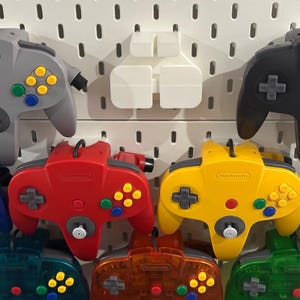 May include: A collection of Nintendo 64 video game controllers in various solid and translucent colours, including grey, black, blue, red, yellow, and green, mounted on a white pegboard wall display with a central white plastic storage bracket.