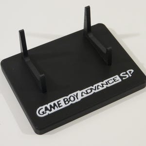 May include: Black Game Boy Advance SP display stand. The stand has a rectangular base with two vertical supports. The words "GAME BOY ADVANCE SP" are printed in white on the base.